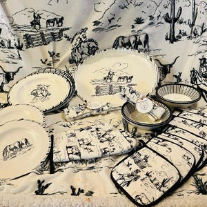 Puede incluir: Un conjunto de vajilla y textiles de cocina con temática del oeste. El juego incluye platos, cuencos, agarraderas y servilletas, todos con ilustraciones en blanco y negro de vaqueros, ganado y paisajes desérticos. Los platos tienen un borde negro.