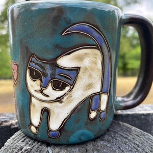 May include: A ceramic mug with a teal glaze and a whimsical cat design. The cat is depicted in white with blue accents and black outlines. The handle is a dark brown color. The mug is likely handmade and suitable for hot beverages.