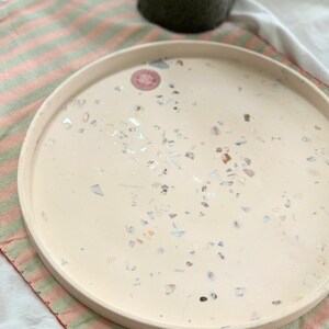 Op de afbeelding: Een ronde, crèmekleurige decoratieve schaal met een gespikkeld ontwerp. De schaal is versierd met kleine, kleurrijke fragmenten en een subtiele, verhoogde rand. De schaal ligt op een roze en groen gestreepte stof.