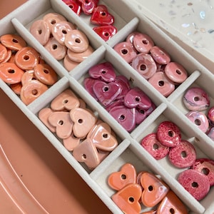 May include: A white box with compartments filled with various heart-shaped ceramic beads in shades of pink, red, and orange. The beads have a glossy finish and are arranged in rows.