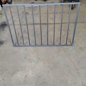 Puede incluir: Una puerta de metal gris con barras verticales. La puerta está sobre un suelo de hormigón.