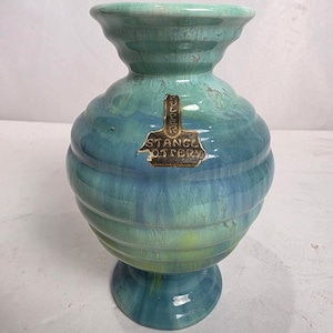 Puede incluir: Un jarrón de cerámica turquesa y azul con un cuerpo bulboso y un borde acampanado. El jarrón tiene una textura acanalada y una base de pedestal. Una etiqueta con el texto "STANCE POTTERY" está adherida al frente.