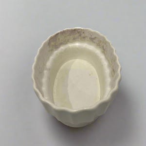Könnte beinhalten: Eine kleine, cremefarbene Keramikschale mit einem gewellten Rand. Das Innere ist ein hellerer Cremeton, und der Rand weist ein subtiles, gealtertes Aussehen auf. Die Schale ist wahrscheinlich zum Servieren oder Ausstellen gedacht.