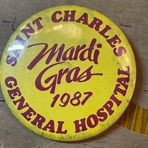 May include: Yellow button with red text that reads "Saint Charles General Hospital Mardi Gras 1987".