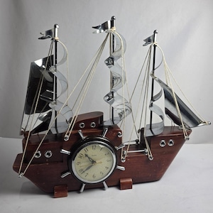 May include: A ship-shaped table clock featuring a dark brown wooden hull, three chrome masts with curved metal sails, and white rigging. A round analog clock face with a ship wheel frame is centered on the side, displaying black numerals.