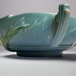May include: A teal ceramic bowl with a textured surface and a raised base. The bowl has a handle on one side and a decorative leaf pattern. The rim has a cream accent.