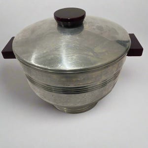 Könnte beinhalten: Ein Vintage-Eiskübel aus Metall mit Deckel und zwei seitlichen Griffen. Der Deckel hat einen dunkelbraunen Knopf. Der Eimer hat horizontale geätzte Linien um die Mitte. Die Griffe sind dunkelbraun.