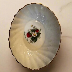 May include: A small, scalloped-edge dish with a gold rim. The interior features a watercolor-style design with a strawberry illustration and the words "Strawberry Ripe". The dish has a gradient of pale yellow and blue.