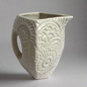May include: A light green ceramic pitcher with a square body and a curved handle. The pitcher features embossed floral and spiral patterns. The spout is angled for easy pouring. The pitcher is a decorative item.