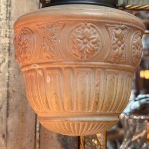 May include: A vintage-style light fixture with a peach-colored glass shade. The shade features a decorative floral and geometric pattern around the top and vertical lines along the bottom. The fixture has a black metal cap.