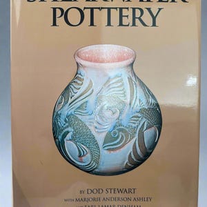 May include: A book cover featuring a Shearwater Pottery vase with a fish design. The vase is blue, white, and brown, with a pink interior. The title "SHEARWATER POTTERY" is at the top, with author and contributor names at the bottom.