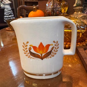 Könnte beinhalten: Cremefarbener Keramikkrug mit Henkel und floralem Design. Das Design zeigt eine orangefarbene und braune Blume mit goldfarbenen Blattakzenten. Ein goldfarbenes Band umrandet den Boden. Der Krug ist wahrscheinlich Vintage oder Retro.