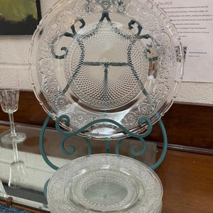 Puede incluir: Platos de vidrio transparente con patrones florales en relieve apilados en un soporte de metal verde azulado. Un plato más grande se muestra arriba. Una pequeña copa de vidrio es visible a la izquierda. Los platos son probablemente para servir o exhibir.