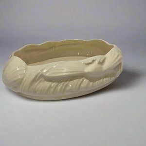 Op de afbeelding: Crèmekleurige keramische plantenbak met een getextureerde, ovale vorm. De plantenbak heeft een gedetailleerd, verhoogd ontwerp rond de randen, dat lijkt op een gestileerd blad of schelp. De binnenkant is glad en de algehele esthetiek is vintage.