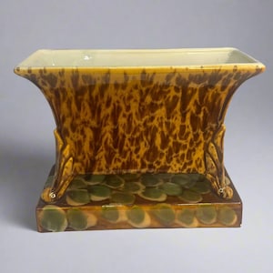 以下が含まれることがあります： 茶色と黄色のまだら模様の釉薬が施された長方形の陶器製のプランター。プランターは緑と茶色の釉薬が施された正方形の土台と、各側に3つの装飾的な足が付いています。