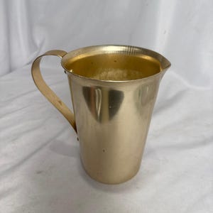 May include: A gold-colored metal pitcher with a handle. The pitcher has a slightly tapered body and a pouring spout. The handle is curved and attached to the side of the pitcher. The interior of the pitcher is also gold-colored.