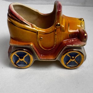 Peut inclure: Un pot de fleurs en céramique en forme de voiture ancienne, dans des tons d'orange, de jaune et de bordeaux. Le pot a des roues bleues et dorées et une poignée. L'intérieur est beige. Design classique et antique.