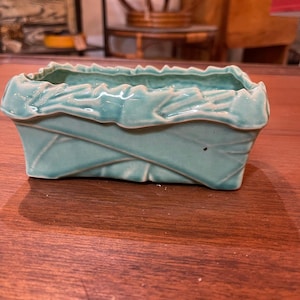 Op de afbeelding: Een tealgroene keramische plantenbak met een geweven ontwerp. De plantenbak heeft een rechthoekige vorm met een licht afgeronde bodem.