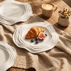 May include: Three white ceramic plates with irregular, wavy, rippled edges. One plate holds a golden-brown croissant, a halved fig, and three fresh blueberries. A beige ceramic mug with latte art and a small pot of dried flowers are also present.