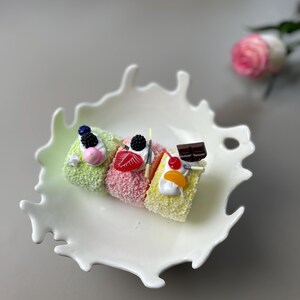 Könnte beinhalten: Drei Mini-Gugelhupf mit unterschiedlich gefärbtem Frosting und Belägen auf einem weißen Teller in Form eines Milchspritzers.