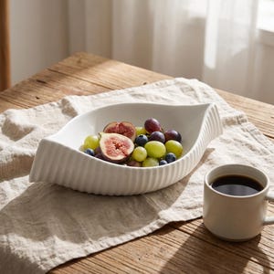 Könnte beinhalten: Eine weiße Keramikschale in Blattform mit gerippter Außenstruktur, gefüllt mit frischen Feigen, grünen Weintrauben und Blaubeeren, neben einer schlichten weißen Keramiktasse mit schwarzem Kaffee auf einer strukturierten Stoffunterlage.