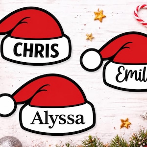May include: Three red and white Santa hat ornaments with black outlines. Each hat features a white brim with a name printed in black text: CHRIS, Emily, and Alyssa. The hats are shaped like traditional holiday headwear with a white pom-pom.