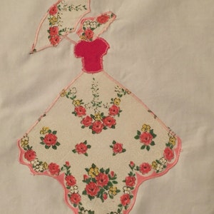 May include: Appliqued handkerchief art depicting a woman with an umbrella. The woman's dress and umbrella are made from floral fabric with red, yellow, and white flowers. The dress has a pink bodice and a scalloped pink border.