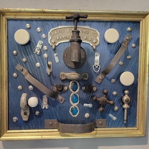 May include: A framed wall art piece featuring a blue wood-grain background with various metal plumbing hardware, including a faucet, handles, and a plaque reading RAPID PLUMBING CO RAPID CITY S.D. accented with white knobs and three blue glass stones.