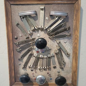Puede incluir: Un marco de madera rectangular que contiene una colección de abridores de botellas de metal vintage dispuestos en un patrón circular sobre un fondo gris texturizado. Tres pomos grandes, dos negros y uno plateado, están en la base.