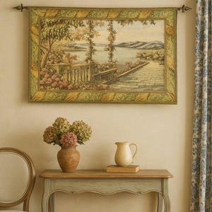 May include: A framed tapestry depicting a scenic landscape with a lake and mountains. The tapestry is framed in green and gold, and hangs above a wooden table with a vase of flowers and a cream-colored pitcher.