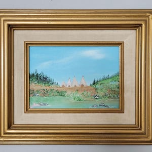 May include: An oil painting depicting a landscape with teepees, a river, and trees under a blue sky. The artwork is framed in a gold-colored, ornate frame, adding a touch of elegance to the scene. The painting evokes a sense of tranquility and natural beauty.