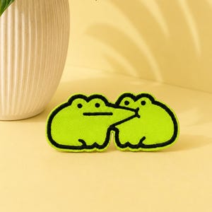 May include: An embroidered patch featuring two lime green frogs with black outlines. The frog on the left has a flat line for a mouth, while the frog on the right has a small circular mouth, appearing as if they are touching or kissing.