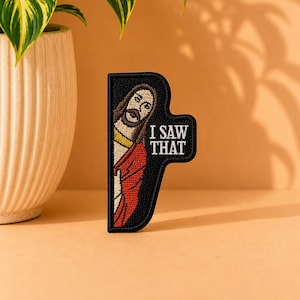 May include: A black embroidered patch featuring a figure with brown hair and a beard wearing a red robe, peeking from the left side. The text "I SAW THAT" is embroidered in white capital letters on the right side of the black background.