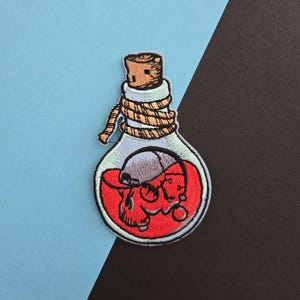 May include: A red and white embroidered patch of a glass bottle with a cork stopper and a skull inside. The bottle is tied with a rope and the skull is filled with red liquid.
