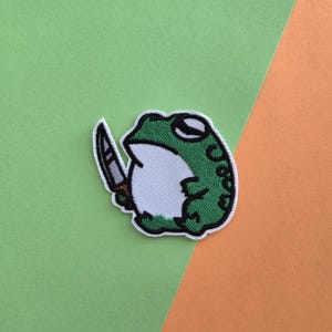 May include: Green frog holding a knife, embroidered patch.