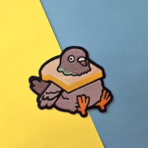 May include: A gray embroidered patch of a pigeon with a yellow and brown sandwich on its head. The patch is on a yellow and blue background.