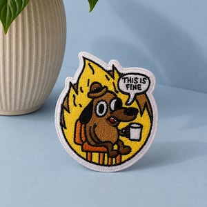 May include: A circular embroidered patch featuring a brown dog sitting on an orange chair amidst yellow flames. The dog holds a white mug, and a white speech bubble above its head contains the black text "THIS IS FINE" in capital letters.
