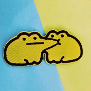 May include: A yellow embroidered patch featuring two cartoon frogs kissing. The frogs have black outlines and details, set against a blue and yellow background. The patch is likely for clothing or accessories.