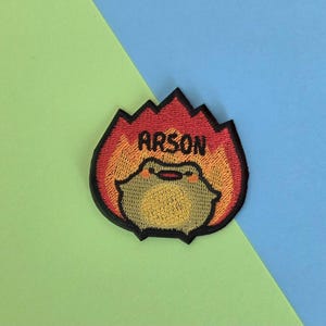 May include: Embroidered patch featuring a cartoon frog in flames. The patch has a red and orange flame design with the word "ARSON" in black letters. The frog has a smiling expression.