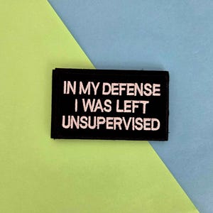 May include: Black rectangular patch with white embroidered text that reads "IN MY DEFENSE I WAS LEFT UNSUPERVISED". The patch is set against a background of lime green and blue.