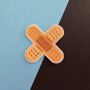 May include: A brown and white fabric patch shaped like a bandage with a white border. The patch is on a blue and black background.