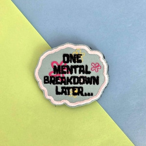 May include: A light gray embroidered patch with the text "ONE MENTAL BREAKDOWN LATER..." in black. The patch has a light pink and yellow floral design. The patch is on a background of light blue and yellow.