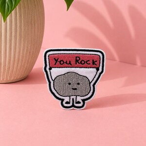 May include: An embroidered patch featuring a grey cartoon rock character with a simple face, holding a pink banner with the black text You Rock. The patch has a white background, a thick black border, and small black embroidered legs at the base.