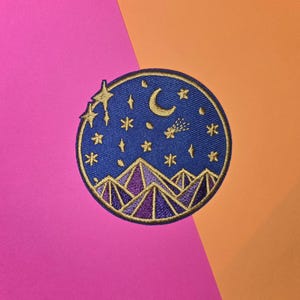 May include: A round, blue embroidered patch with a gold outline. The patch features a crescent moon, a shooting star, and a mountain range with purple and gold accents against a dark blue background with gold stars.