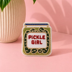 May include: An embroidered patch shaped like a glass jar filled with green pickles. The jar features a white rectangular label in the center with the red text PICKLE GIRL. The lid is blue, and the entire patch has a white border outline.