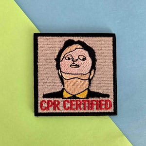 May include: Embroidered patch featuring a cartoon portrait with a light skin tone, black hair, and glasses. The character is wearing a black suit with a yellow collar. The text "CPR CERTIFIED" is in red. The patch has a black border.