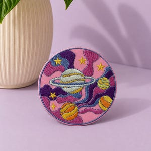 May include: A circular embroidered patch featuring a space theme with a central planet with rings, two smaller spherical planets, and several yellow stars. The design incorporates swirling patterns in shades of pink, purple, and blue.