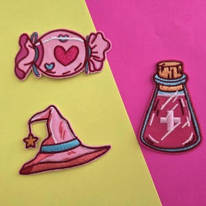 May include: Three embroidered iron-on patches: a pink candy with a heart, a pink witch's hat with a star, and a pink potion bottle with a white cross.