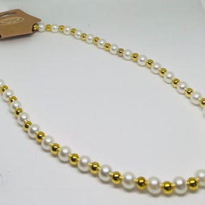 May include: A necklace with alternating white and gold beads. The beads are strung together in a continuous loop. The round beads are evenly spaced. A tag is attached at the top of the necklace.