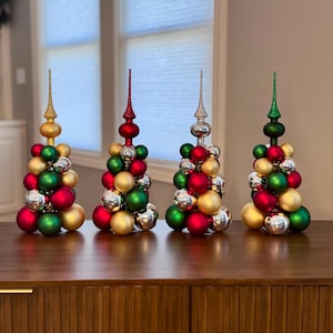 May include: Four decorative Christmas tree ornaments. Each tree is made of red, green, gold, and silver ornaments. The trees have a decorative spire at the top. The trees are displayed on a wooden surface.